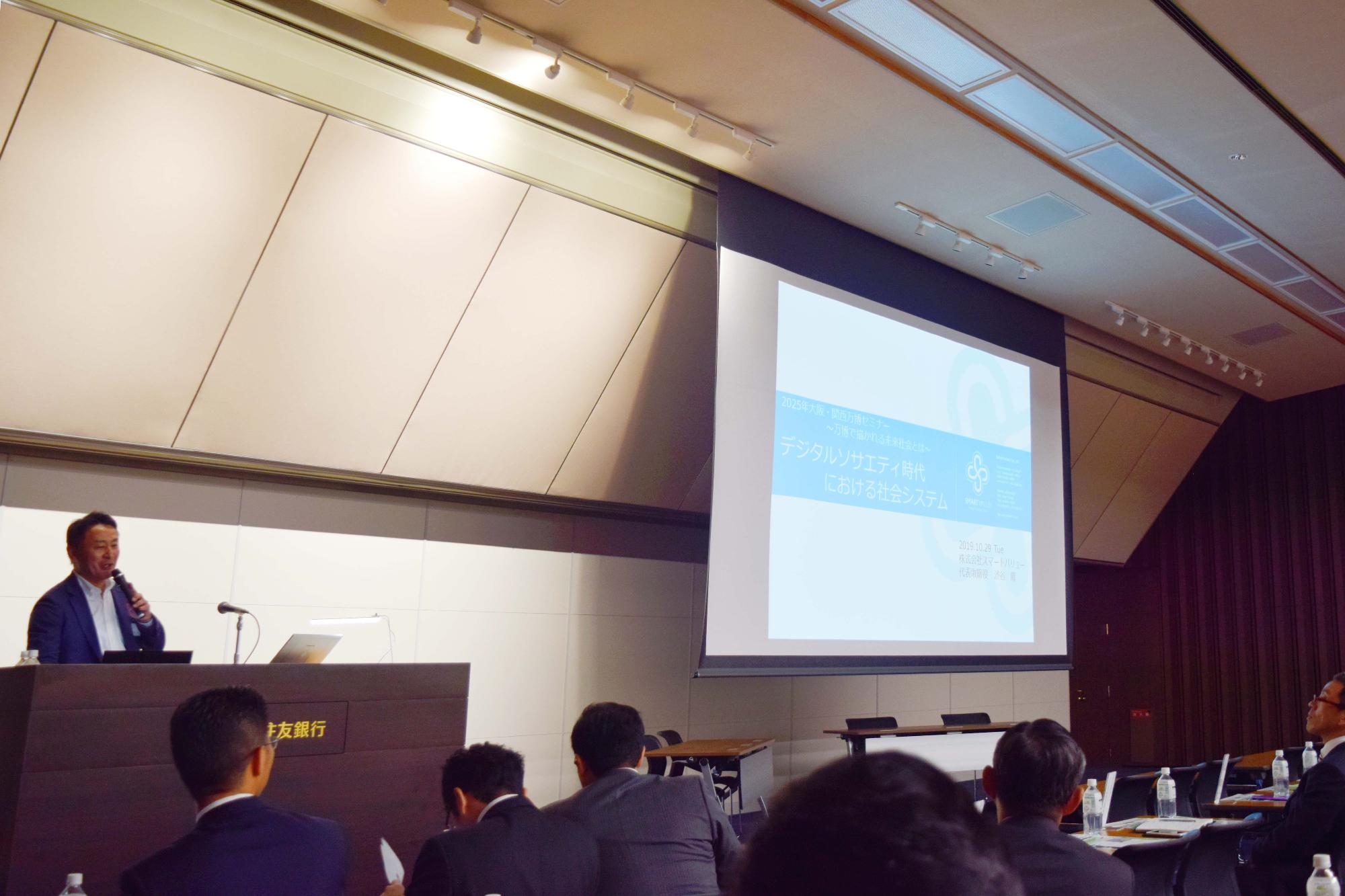 プロジェクターで映し出されたスライドの左側で、演説台にてマイクを持ちながら話している男性の写真と、話を聞く数名の男性の写真