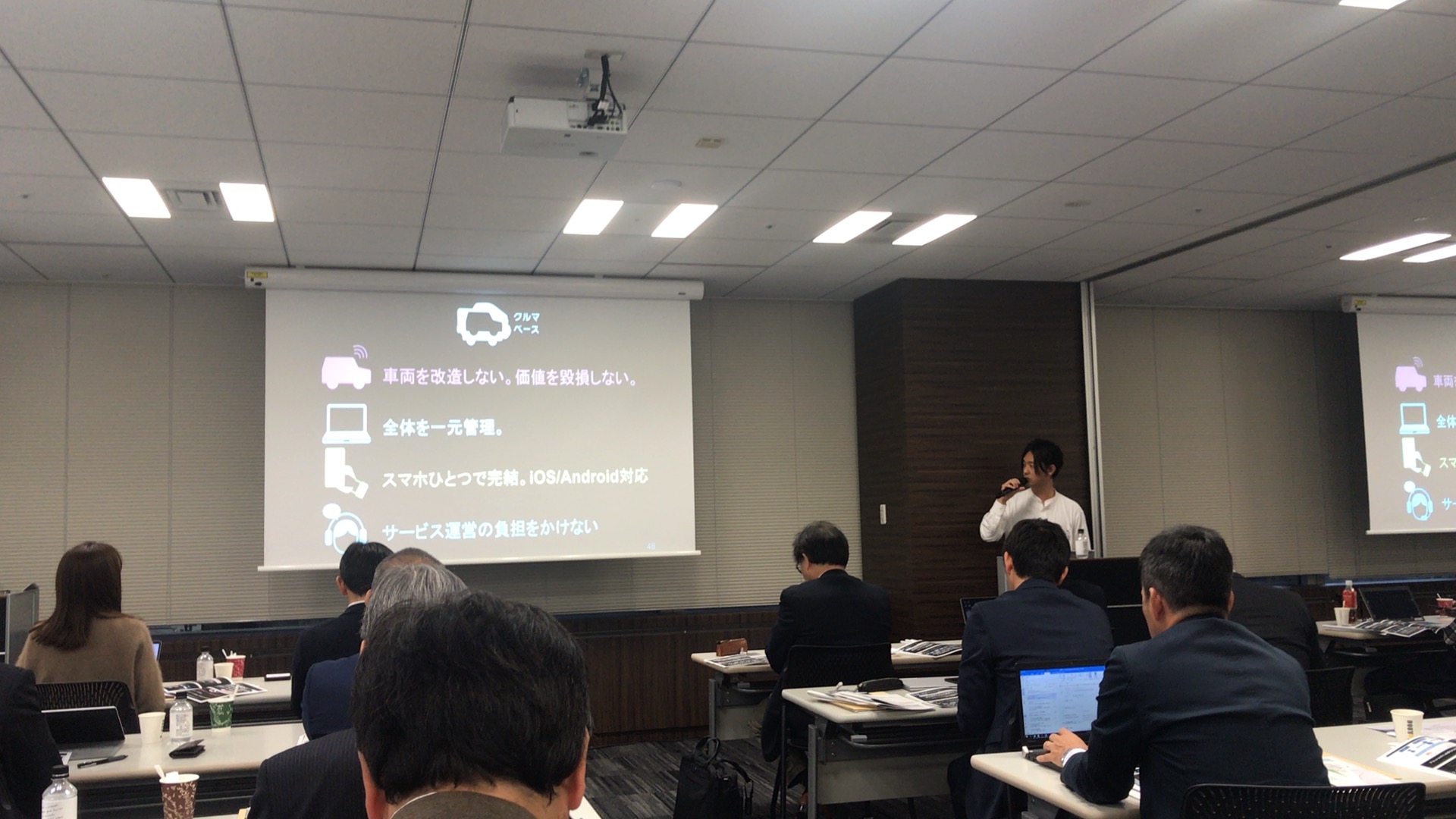 プロジェクターに映し出されたスライドを見ながらマイクをもって部屋の前で話す男性と、机と椅子に座って話を聞く男女数名の写真。スライドには「クルマベース 車両を改造しない。価値を毀損しない。 全体を一元管理。 スマホをひとつで完結。IOS/Android対応 サービス運営の負担をかけない」と書かれている。