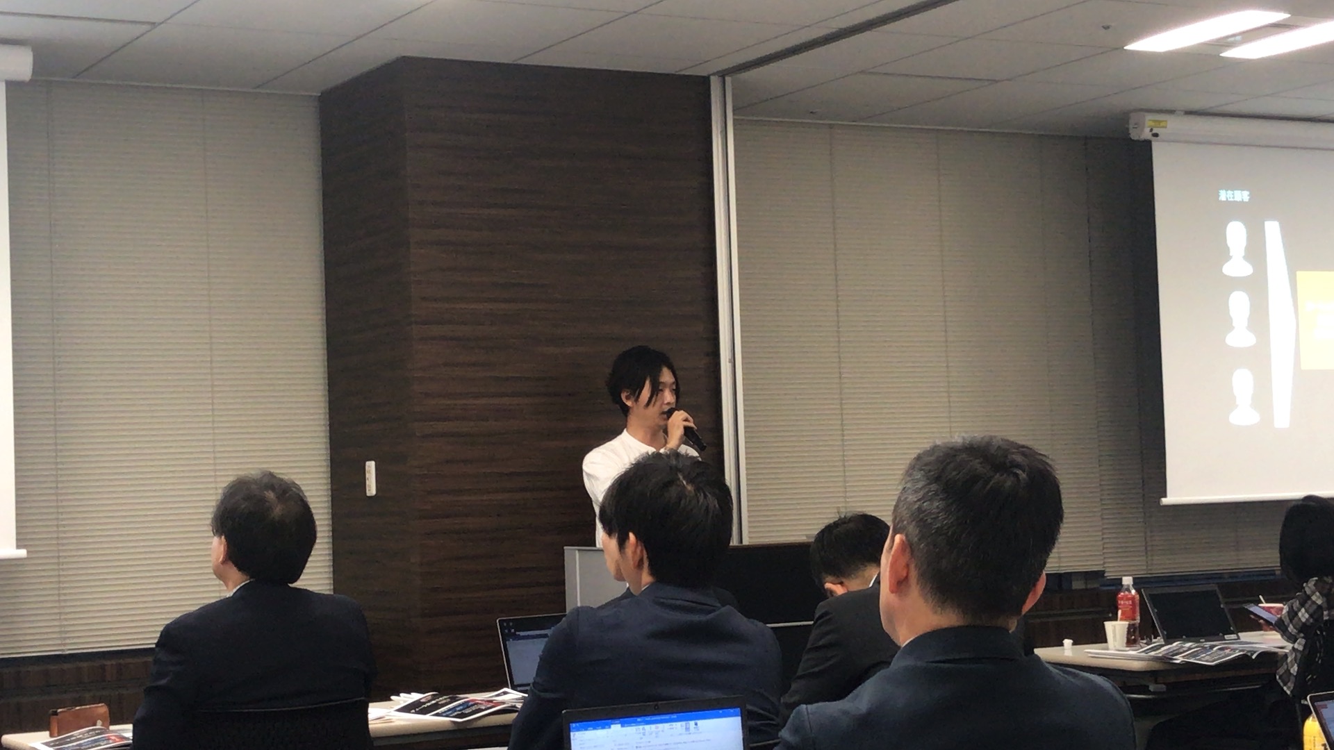部屋の前でマイクを持って傍聴者を見ながら話す男性の写真と、机と椅子に座りながら話を聞く数名の大人達の写真