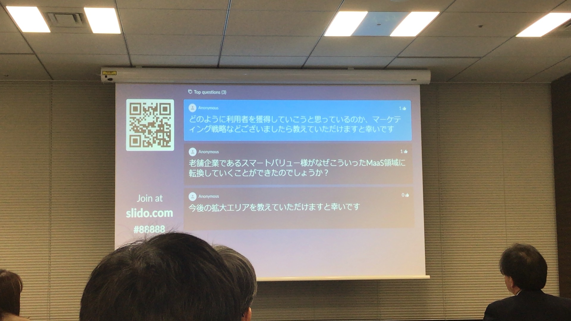 プロジェクターで映し出されたスライドを移した画像。スライドには「どのように利用者を獲得していこうと思っているのか、マーケティング戦略などございましたら教えていただけますと幸いです。 老舗企業であるスマートバリュー様がなぜこういったMaaS領域に転換していくことができたのでしょうか？ 今後の拡大エリアを教えていただけますと幸いです」と書かれている。