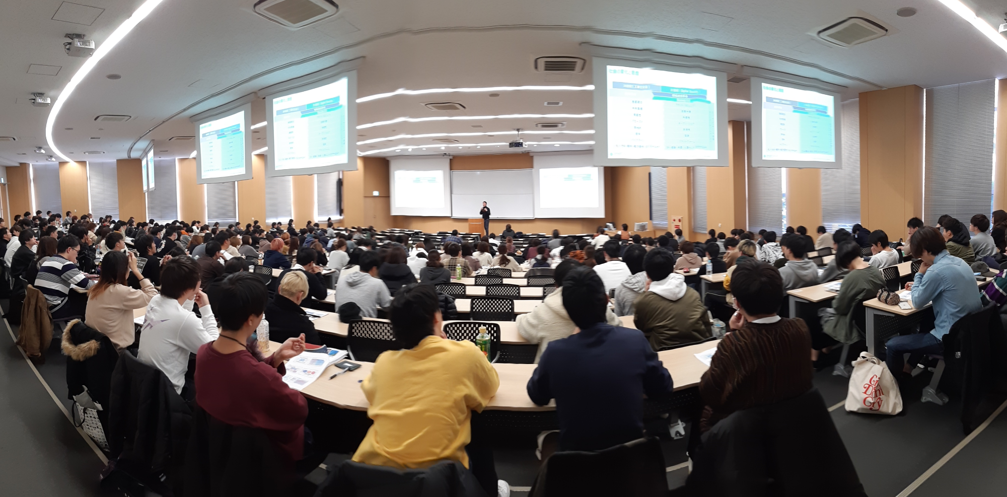 大学の講義室で壇上にて講演を行う一人の男性と、構成を聞く多数の学生の画像