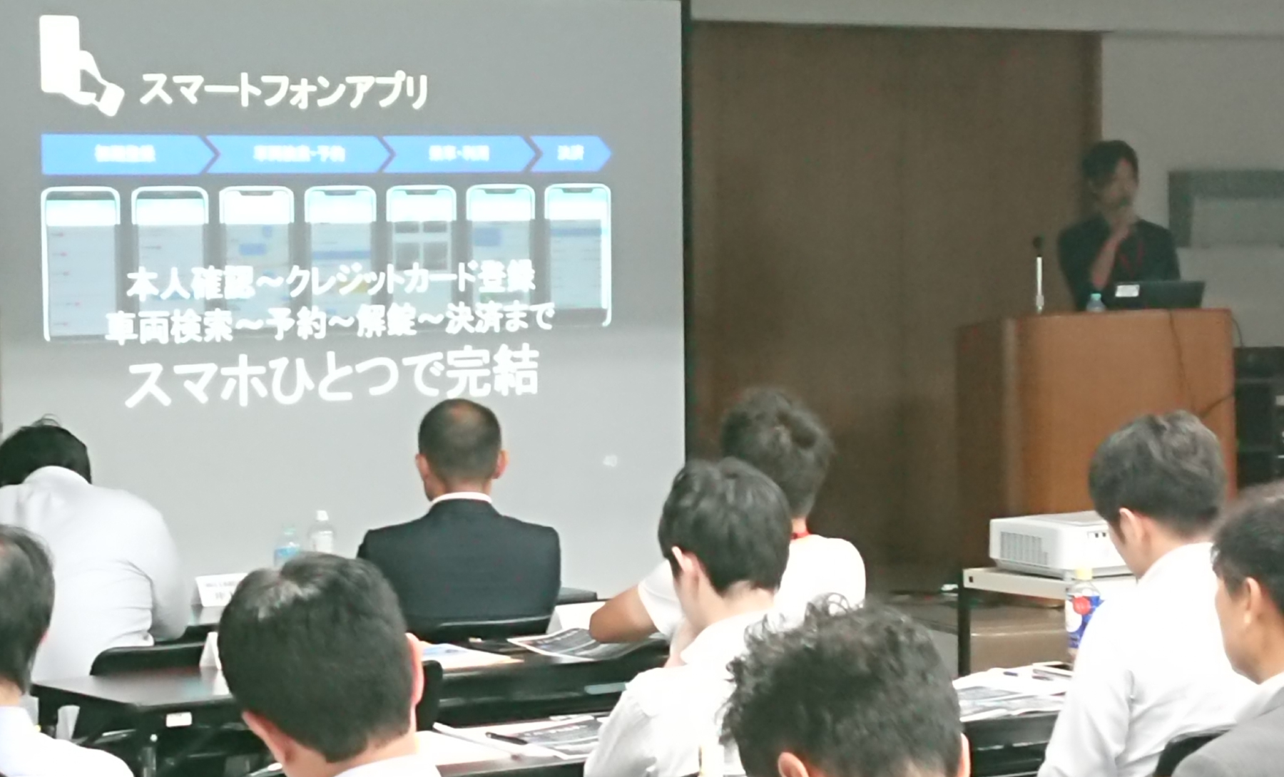 講演台にてノートパソコンを開きながらマイクをもって部屋の前で話をしている男性と、机と椅子に座って話を聞く数名の男性の写真。また話をしている男性の左側にプロジェクターから映し出されたスライドがあり、スライドには「本人確認～クレジットカード登録 車両検索～予約～解錠～決済まで スマホひとつで完結」と書かれている。