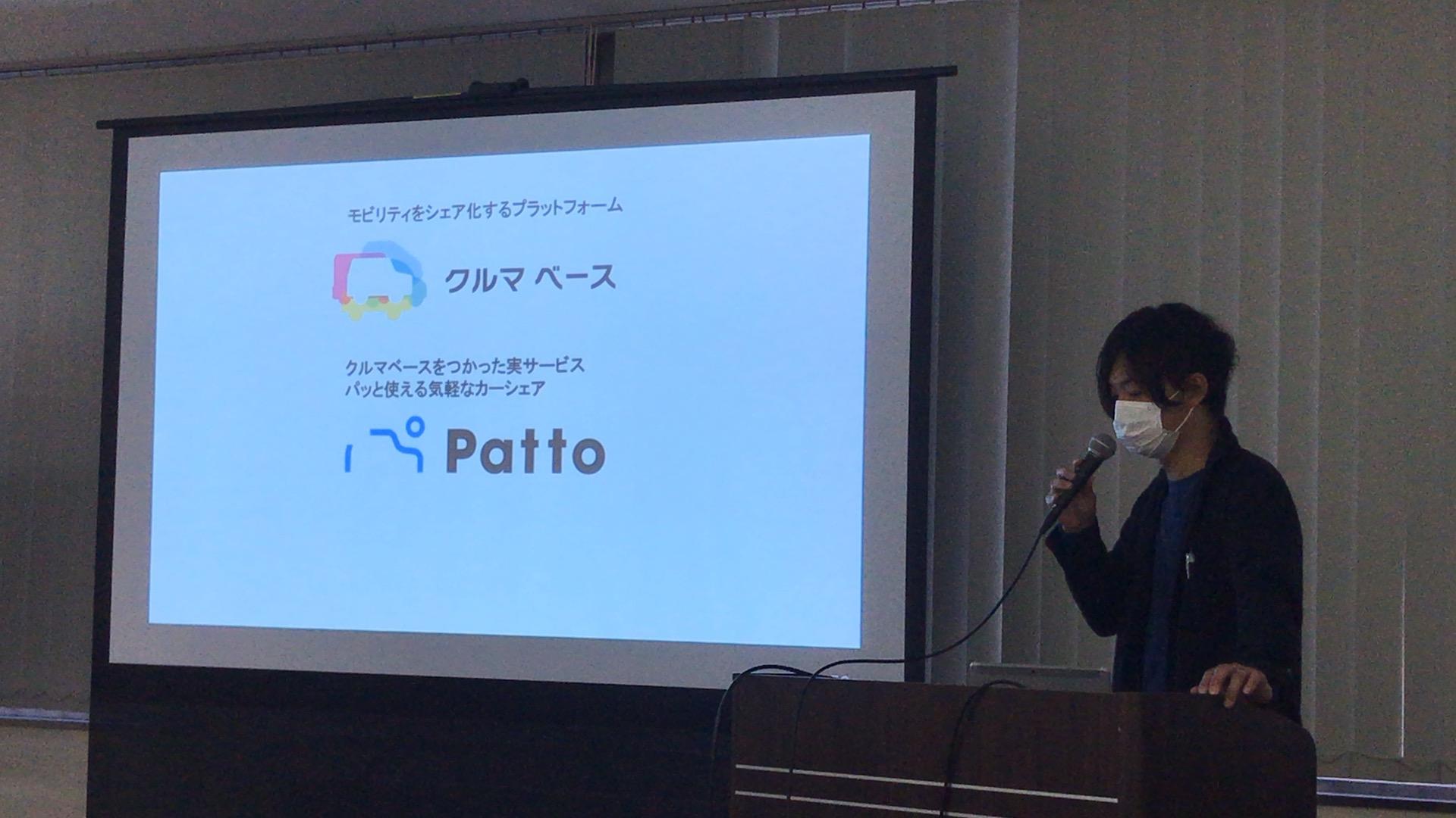 男性がプロジェクターで映し出されたスライドの右斜め前で、スライドを見ながらマイクを持って話している画像。スライドには「Patto 3つの特徴 1.パッと乗れる！地域密着カーシェア！ 2.カーシェアはつ！やさしい運転でおトクに利用！ 3.スマホでクルマの予約から鍵の解錠・施錠まで！」と書かれている。
