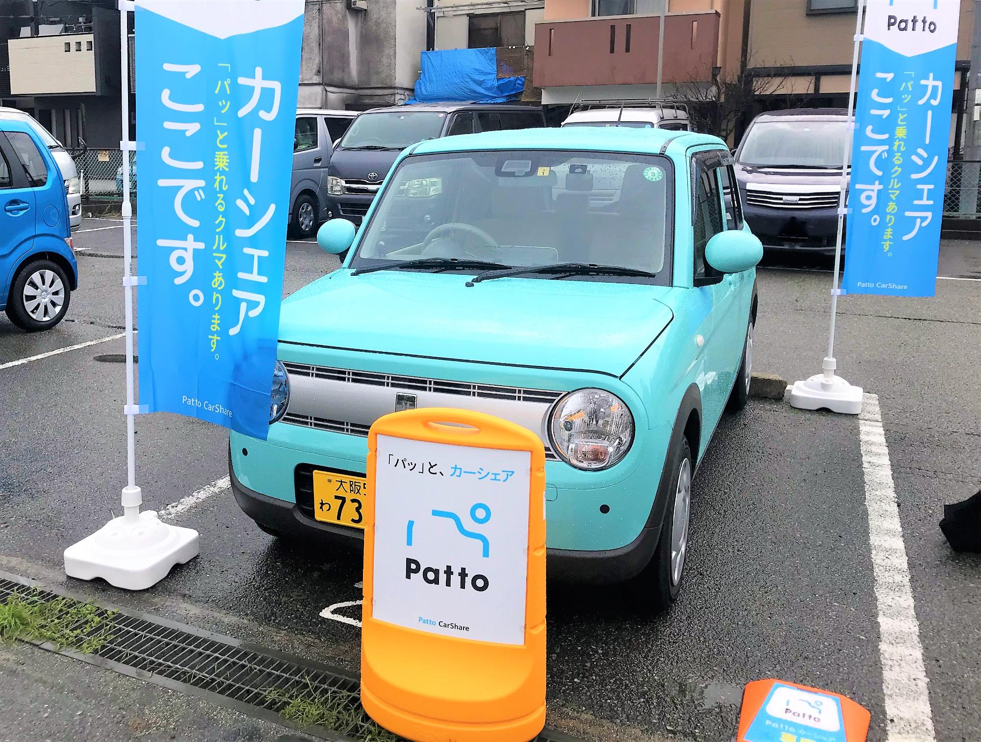 駐車場に駐車されている水色の軽自動車の画像。軽自動車の正面には「パッと、カーシェア Patto」と書かれたカラーコーンが、軽自動車の左斜め前と右斜め後ろには「カーシェア パッと乗れるクルマあります。ここです。」と書かれた旗が立っている。