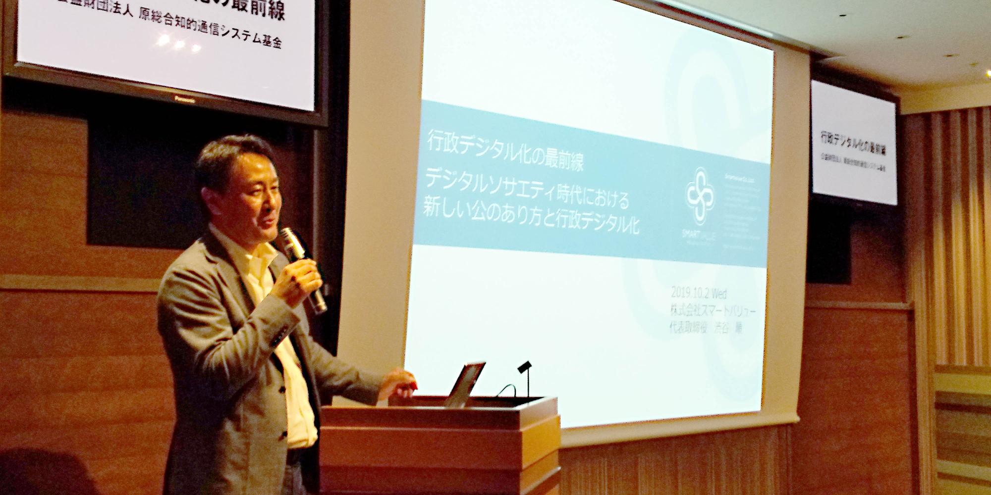 写真の左手前にマイクをもった男性が講演台で話をしており、写真右奥にはプロジェクターに映し出されたスライドが写っている写真。スライドには「行政デジタルの最前線 デジタルソサエティ時代における新しい公のあり方と行政デジタル化」と書かれている。