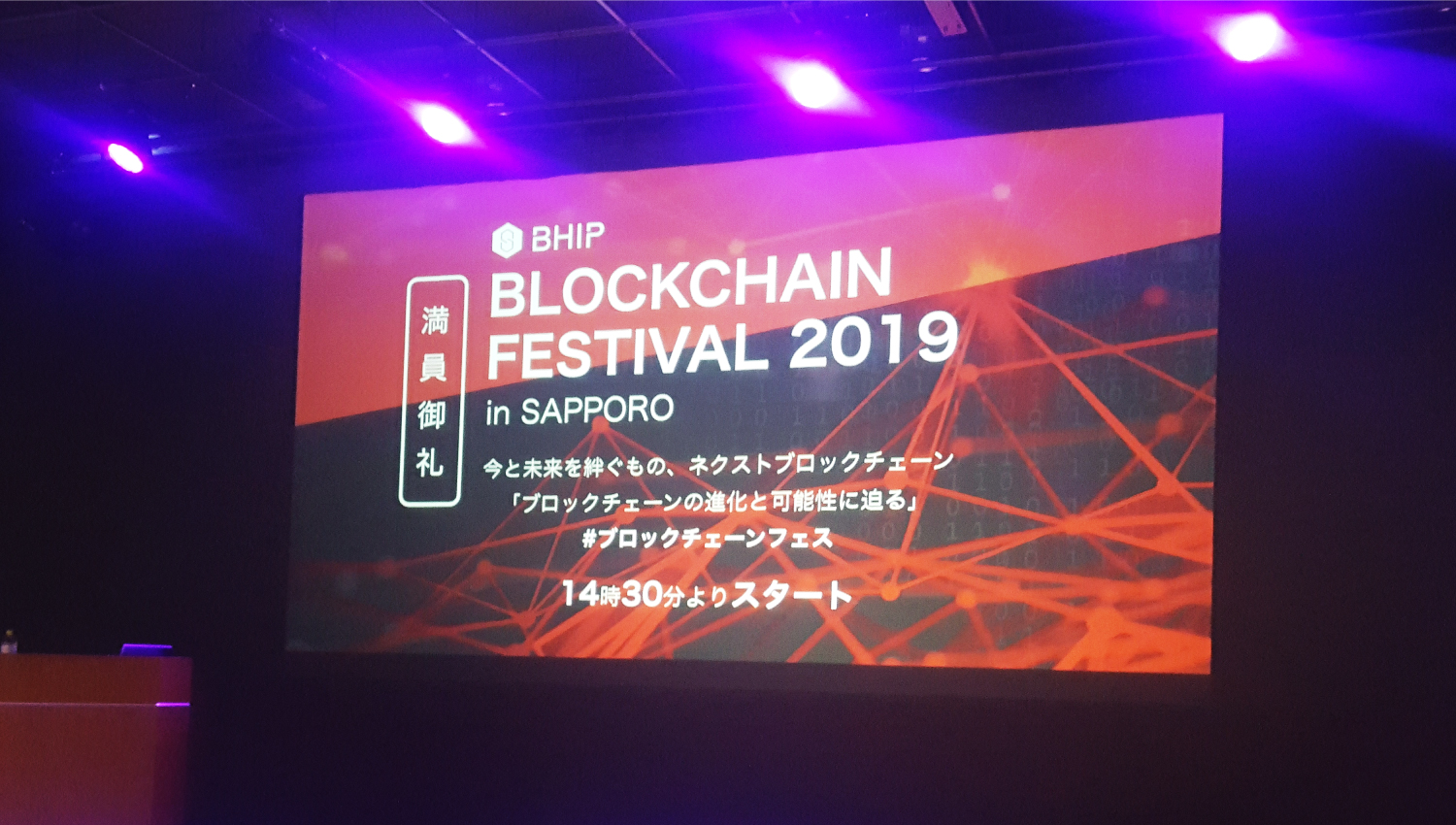 プロジェクターで映し出されたスライドを撮影した写真。スライドには「BLOCKCHAIN FESTIVAL 2019 in SAPPORO 今と未来を絆ぐもの、ネクストブロックチェーン ブロックチェーンの新化と可能性に迫る #ブロックチェーンフェス 14時30分よりスタート」と書かれている。