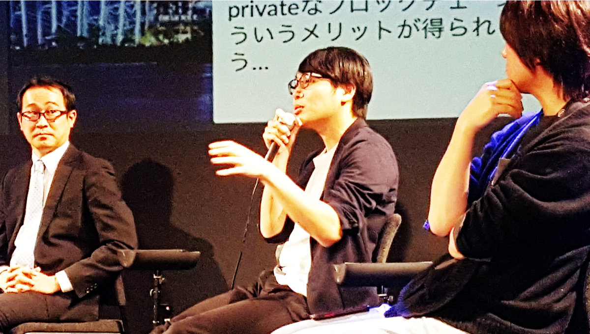 椅子に座りながらスポットライトの光が当たった3名の男性が写っており、そのうち中央の男性が右手にマイクをもって、左手で手振りを行いながら話をしている写真