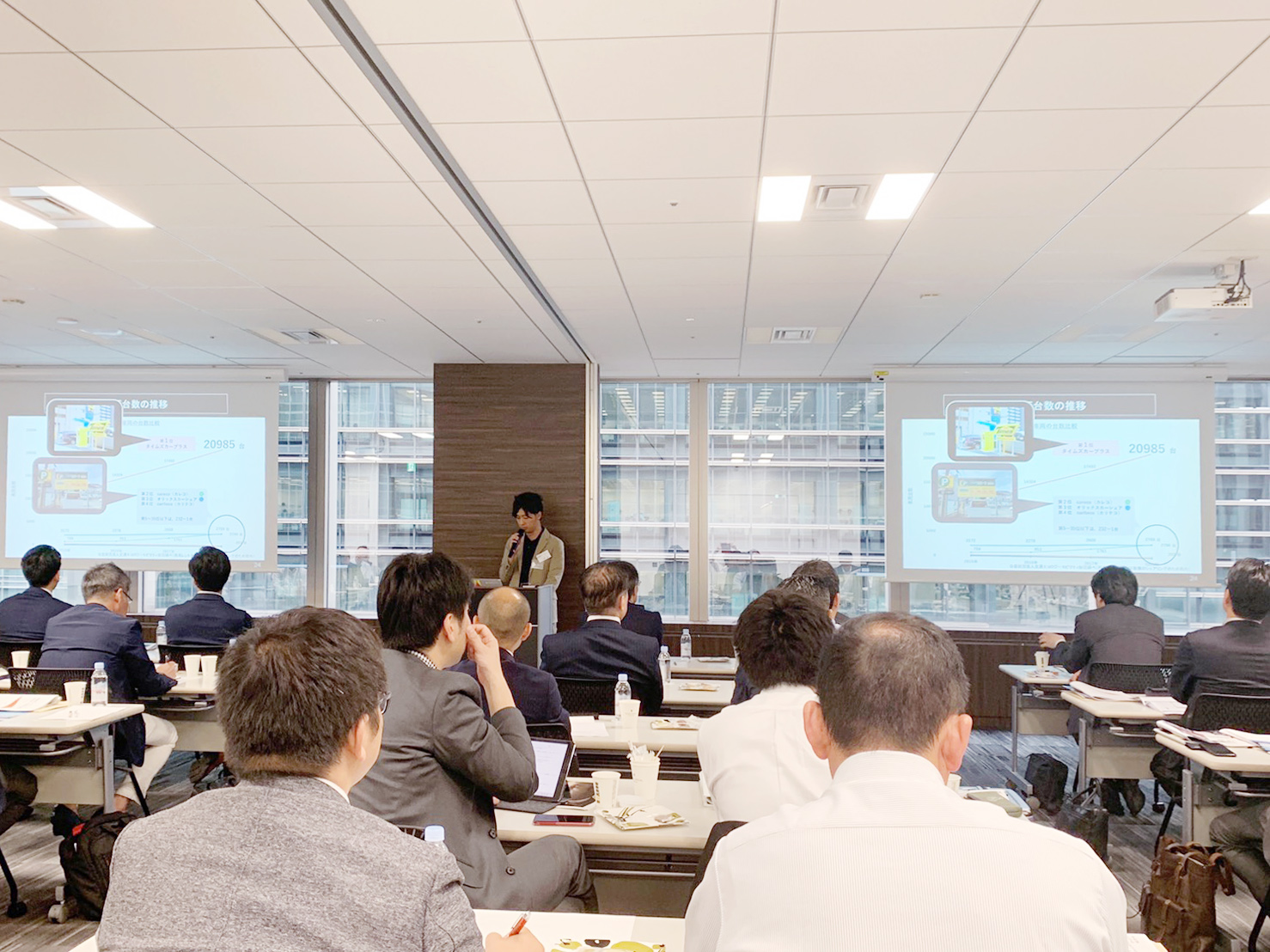 部屋の前で、講演台にてマイクをもちながら話をしている男性と、机と椅子に座って話を聞く十数名の大人の写真。話をしている男性の両隣にはプロジェクターから映し出されたスライドがあり、スライドには折れ線グラフと写真が掲載されている。